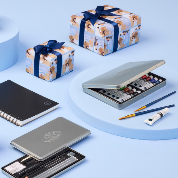 A flat lay arrangement featuring two wrapped gifts with blue bows, a light blue circular platform, a gray art supply case with paints and brushes, a white tube, a blue pen, and a spiral notebook. The background is light blue.