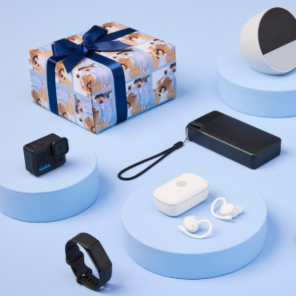 A neatly arranged flat lay featuring a gift-wrapped box, a camera, wireless earbuds, a portable charger, a tracking device, and a fitness tracker, all on light blue circular platforms against a blue background.