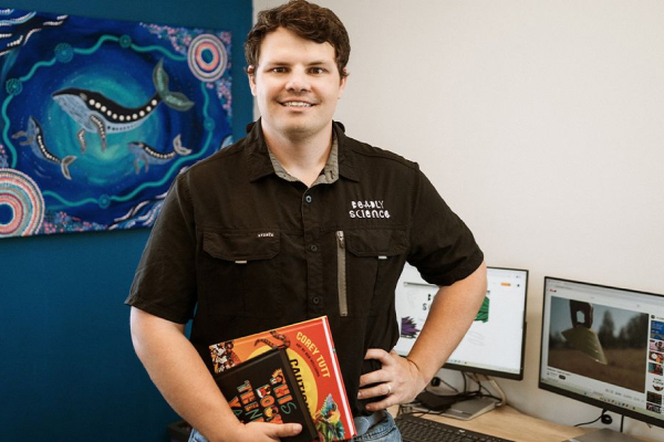 A person standing in an office setting holding books, with computers and artwork in the background.