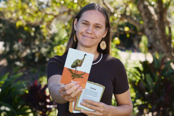 A woman is smiling while holding a card with an illustration of an emu on it, standing outdoors among greenery.