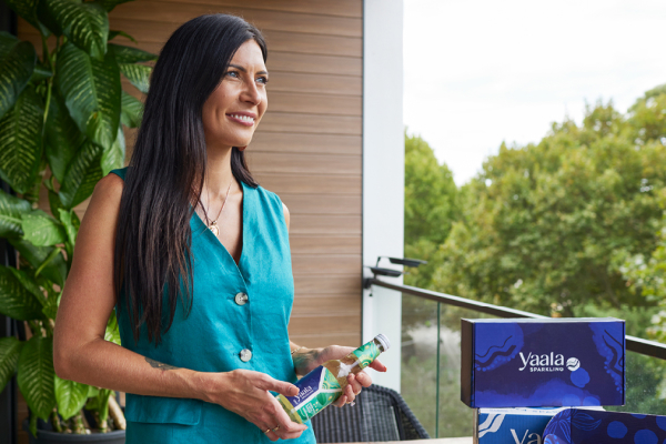 A woman with long dark hair stands on a balcony holding a bottle, wearing a teal outfit, with plants and a box labeled 'yaala' in the background.